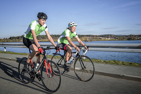 Zwei Rennradfahrer in voller Fahrt vor einem See und blauem Himmel